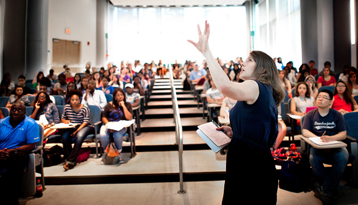 An instructor gestures to an off-screen presentation while a large room of students listen.